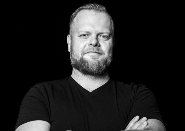 Vīrietis ar bārdu melnā fonā, skatās tieši kamerā un smaida viegli.